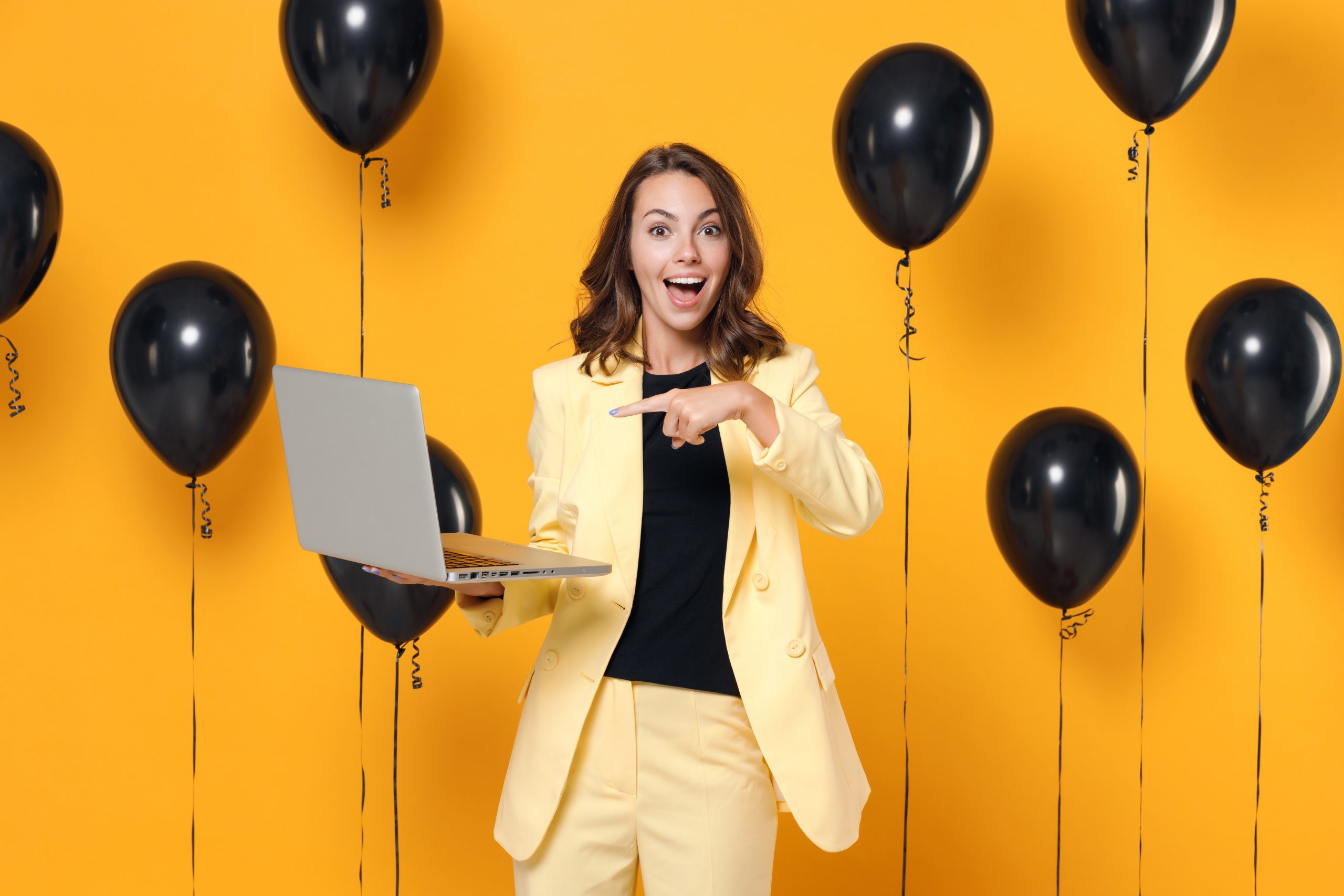 Excited amazed young brunette woman 20s wearing basic light suit jacket pointing index finger on laptop pc computer on yellow colour background with air balloons studio portrait. Black friday sale