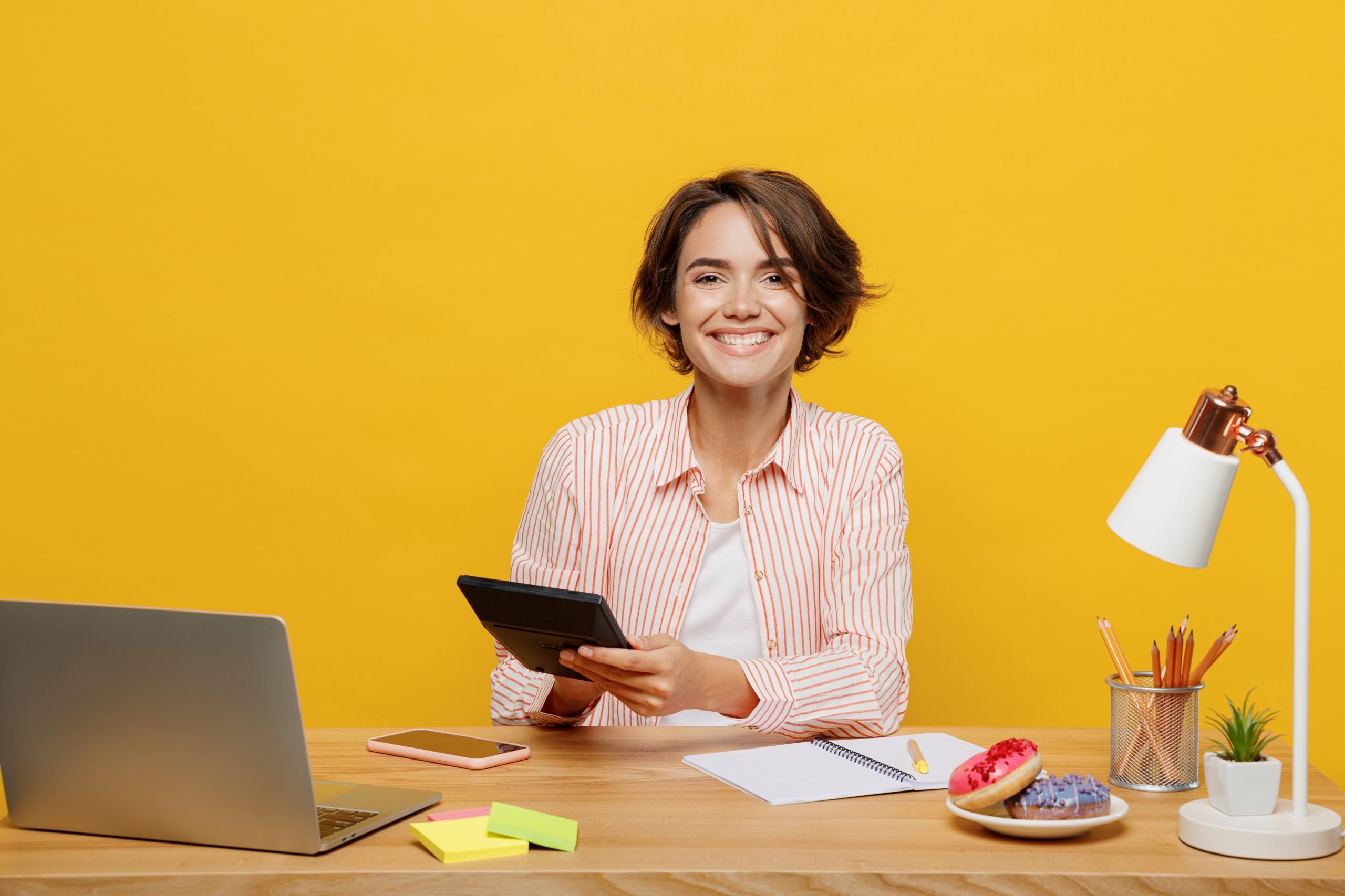 Young fun successful happy employee business woman wear casual shirt sit work at office desk with pc laptop use calculator write isolated on plain yellow color background. Achievement career concept